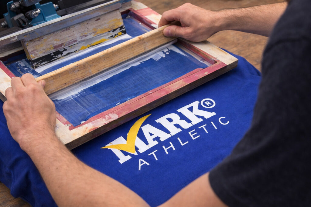 Proceso de serigrafía aplicando logo de Mark Athletic sobre playera azul para estampado textil